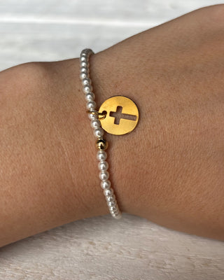 Pearl bracelet with a gold disc featuring a cross on a wrist against a neutral background