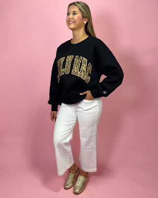 Woman wearing a black sweatshirt with gold text, white pants, and gold shoes on a pink background