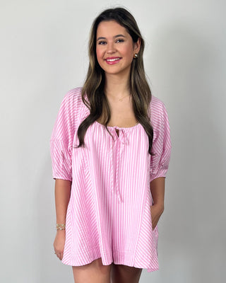 Smiling woman posing against a neutral backdrop wearing a pink striped flowy romper with puff sleeves, a scoop neckline with a dainty tie detail, and roomy front pockets, finished with clear slide sandals and delicate gold jewelry.