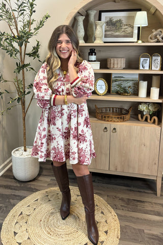 Woman in a floral dress and knee-high boots standing in a room with a wooden cabinet and decorative items.
