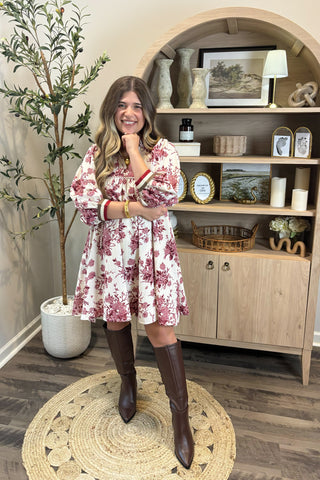 Woman in a floral dress and knee-high boots standing in a room with wooden shelves and decor.