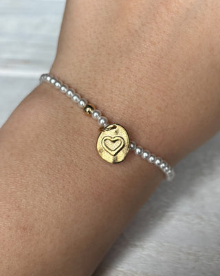 Gold heart charm on a pearl bracelet on a wrist against a white background