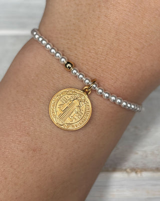 Gold coin charm on a pearl bracelet on a wrist against a neutral background