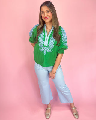 Woman wearing a green embroidered top and light blue jeans against a pink background