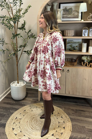 Woman in a floral dress standing in a room with wooden flooring and a plant.