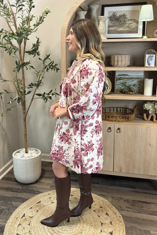 Woman in a floral dress standing in a room with a wooden cabinet and decorative items.