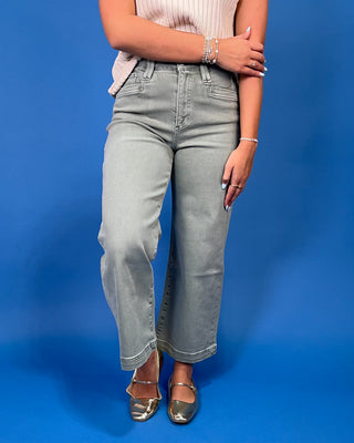 A model wearing light gray cropped jeans with a side slit, paired with a pink ribbed top and metallic gold flats, standing against a solid blue background.