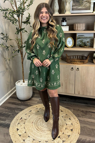 Woman in a green dress with a pattern standing in a room with wooden flooring and a plant.