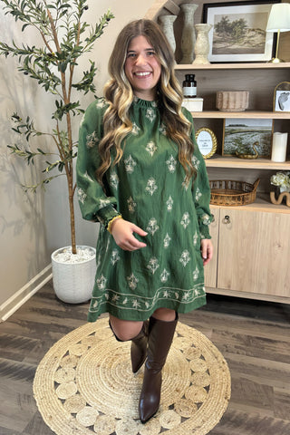 Woman in a green dress standing in a room with wooden flooring and a bookshelf.