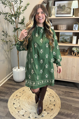 Woman wearing a green patterned dress standing in a room with wooden flooring and a plant.