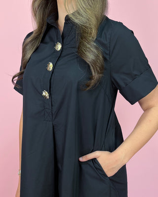 Person wearing a black dress with gold buttons against a pink background