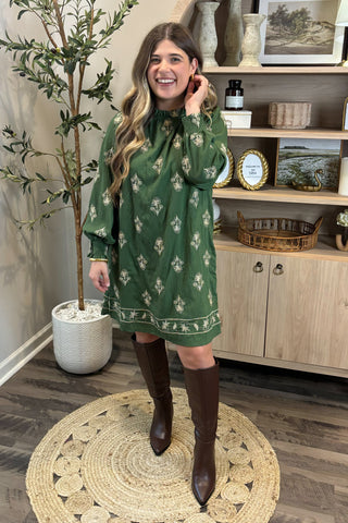 Woman in a green dress with white patterns standing in a room with a plant and shelves.