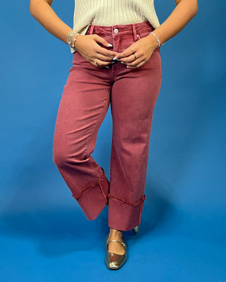 A person wearing dusty rose wide-leg jeans with frayed rolled hems, paired with a cream ribbed top and metallic flats, standing against a solid blue background.