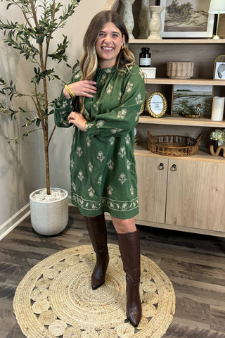 Woman in a green dress with a pattern, standing in a room with wooden flooring and a plant.