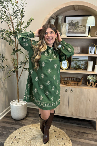 Woman in a green dress with floral pattern standing in a room with wooden shelves and decor.
