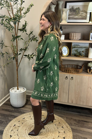 Woman in a green dress standing in a room with wooden shelves and decor.