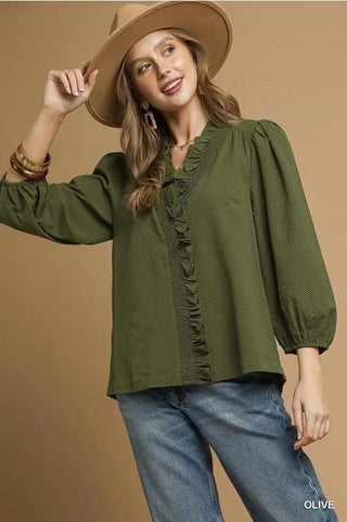 Woman wearing a green blouse, blue jeans, and a beige hat against a brown background