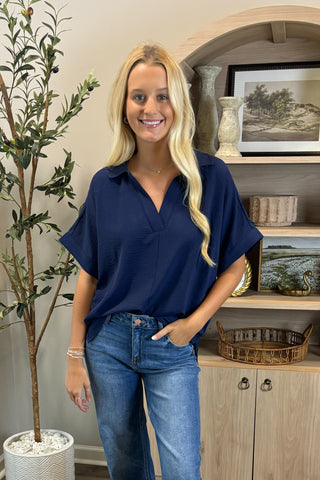 Woman wearing a navy blue shirt and jeans standing in a room with a plant and shelves.