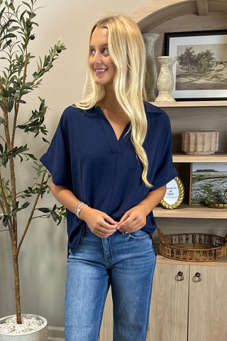 Woman wearing a navy blue shirt and jeans standing in a room with a plant and shelves.