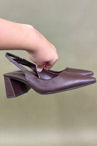 Brown high-heeled shoes held by a hand against a neutral background