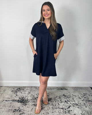 Smiling woman standing against a neutral background wearing a classic black shift dress with a relaxed fit, V-neckline, and striped cuff details on the sleeves, styled with clear slide sandals.