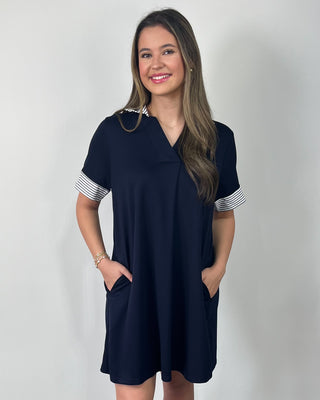 Smiling woman standing against a neutral background wearing a classic black shift dress with a relaxed fit, V-neckline, and striped cuff details on the sleeves, styled with clear slide sandals.
