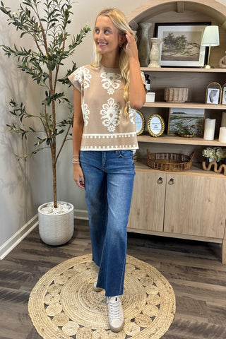 Woman wearing a lace top and blue jeans standing in a room with a plant and wooden cabinet.
