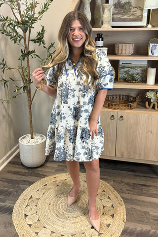 Woman in a floral dress standing in a room with a plant and shelves.