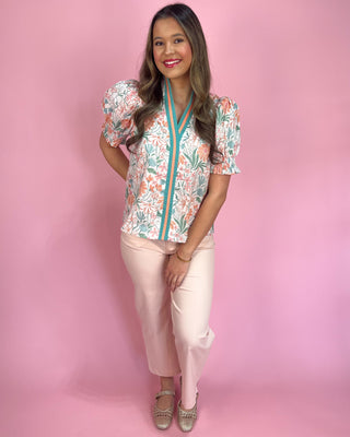 Woman wearing a floral blouse with a zipper and light pink pants on a pink background