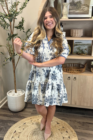 Woman in a floral dress standing in a room with a plant and shelves.