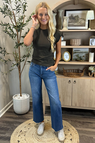 Woman wearing a black t-shirt and blue jeans standing in a room with a plant and shelves.
