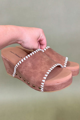 Brown suede clog shoe with white stitching held by a hand against a plain background
