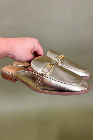 Gold loafers with a gold buckle held by a hand against a beige background
