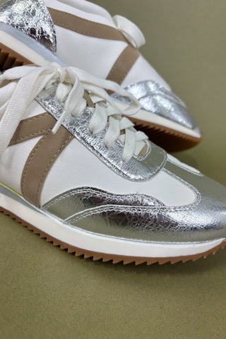 Silver and white sneakers with a hand holding them against a beige background