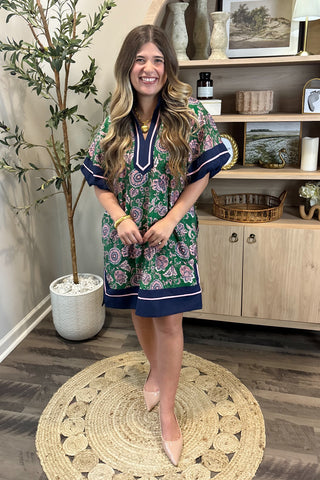 Woman in a floral dress standing in a room with wooden flooring and decor.