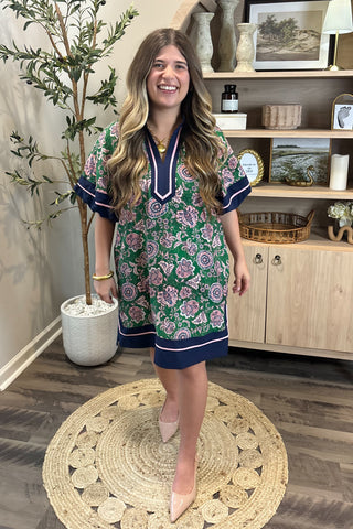 Woman in a floral dress standing in a room with a plant and wooden cabinet.