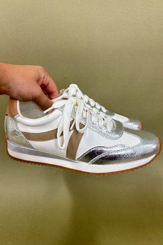 Silver and white sneakers with a hand holding one shoe against a beige background