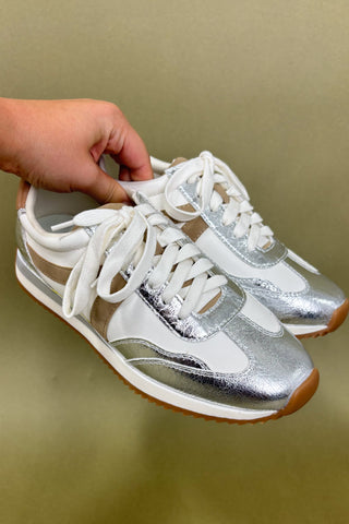Silver and white sneakers with a hand holding them against a beige background
