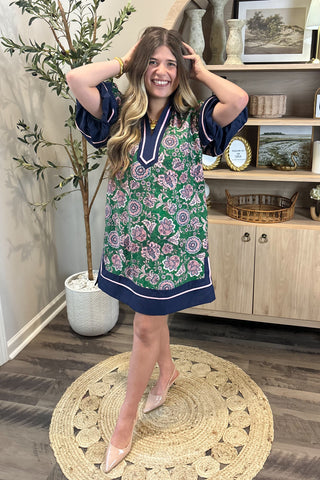 Woman in a floral dress standing in a room with a plant and wooden shelf.