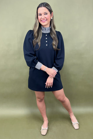 Woman wearing a navy dress with a striped shirt underneath against a green background