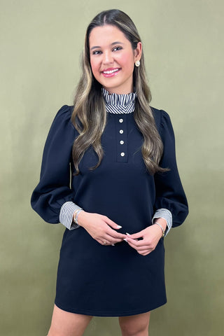 Woman wearing a navy dress with a striped shirt underneath against a green background