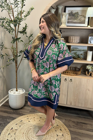 Woman in a floral dress standing in a room with a plant and wooden cabinet.
