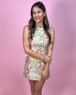 Pink and green floral sleeveless mini dress with a high neckline and structured fit, featuring a vibrant botanical print; styled with simple jewelry against a pink backdrop.