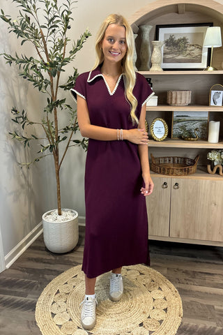 Woman in a purple dress standing in a room with a plant and shelves.