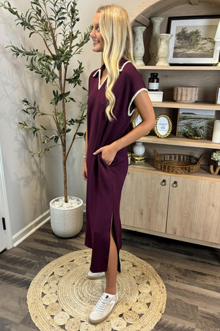 Woman in a purple dress standing in a room with a plant and wooden cabinet.