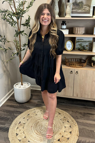 Woman in a black dress standing in a room with a plant and wooden cabinet.