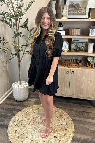 Woman in a black dress standing in a room with a plant and shelves.