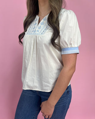 Woman wearing a white blouse with blue embroidery and cuffs against a pink background