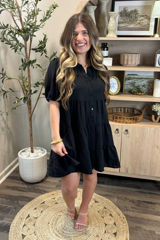 Woman in a black dress standing in a room with a plant and wooden cabinet.