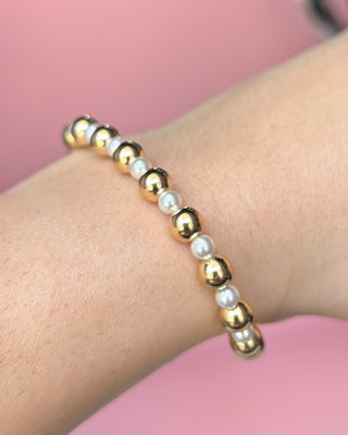 Gold and pearl beaded bracelet on a wrist with a pink background